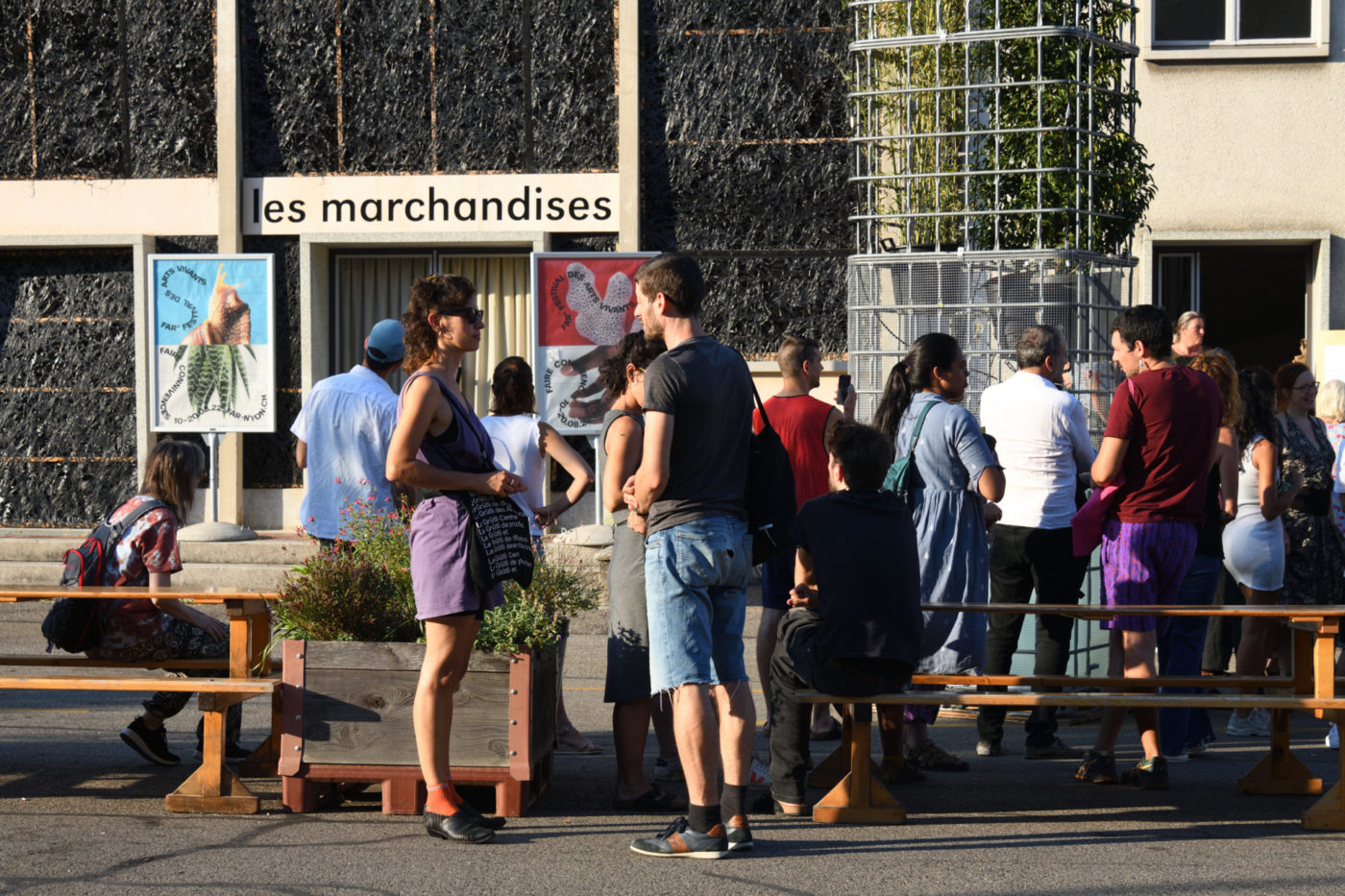Des personnes se trouvent dans la cour devant la salle des Marchandises. On voit les affiches de l'édition 2022, Faire connivences.