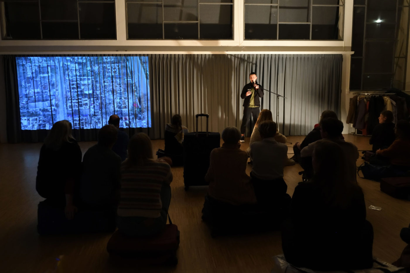We see an artist talking into a microphone in a theater room. Before him are sat people from the public. In the back next to the artist is projected a blurred image.