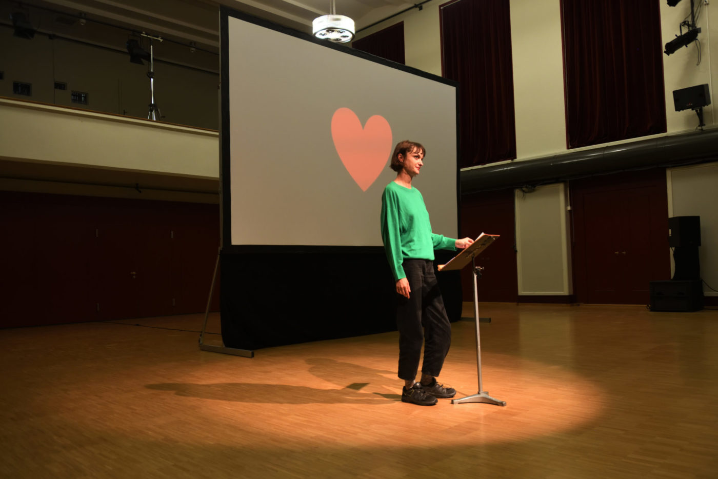 The artist Marion Thomas stands on a scene. Behind her is projected a red heart on a black background.