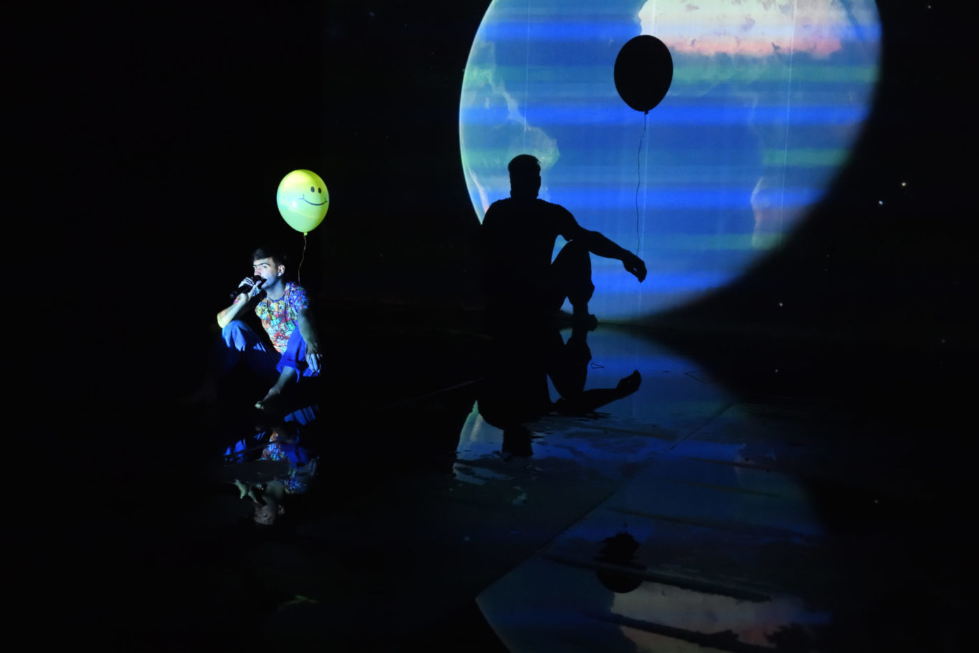 The artist Savino Caruso holds a balloon in his back, that floats. His shadow is reflected in the back in a projection of Planet Earth. He talks into a microphone.