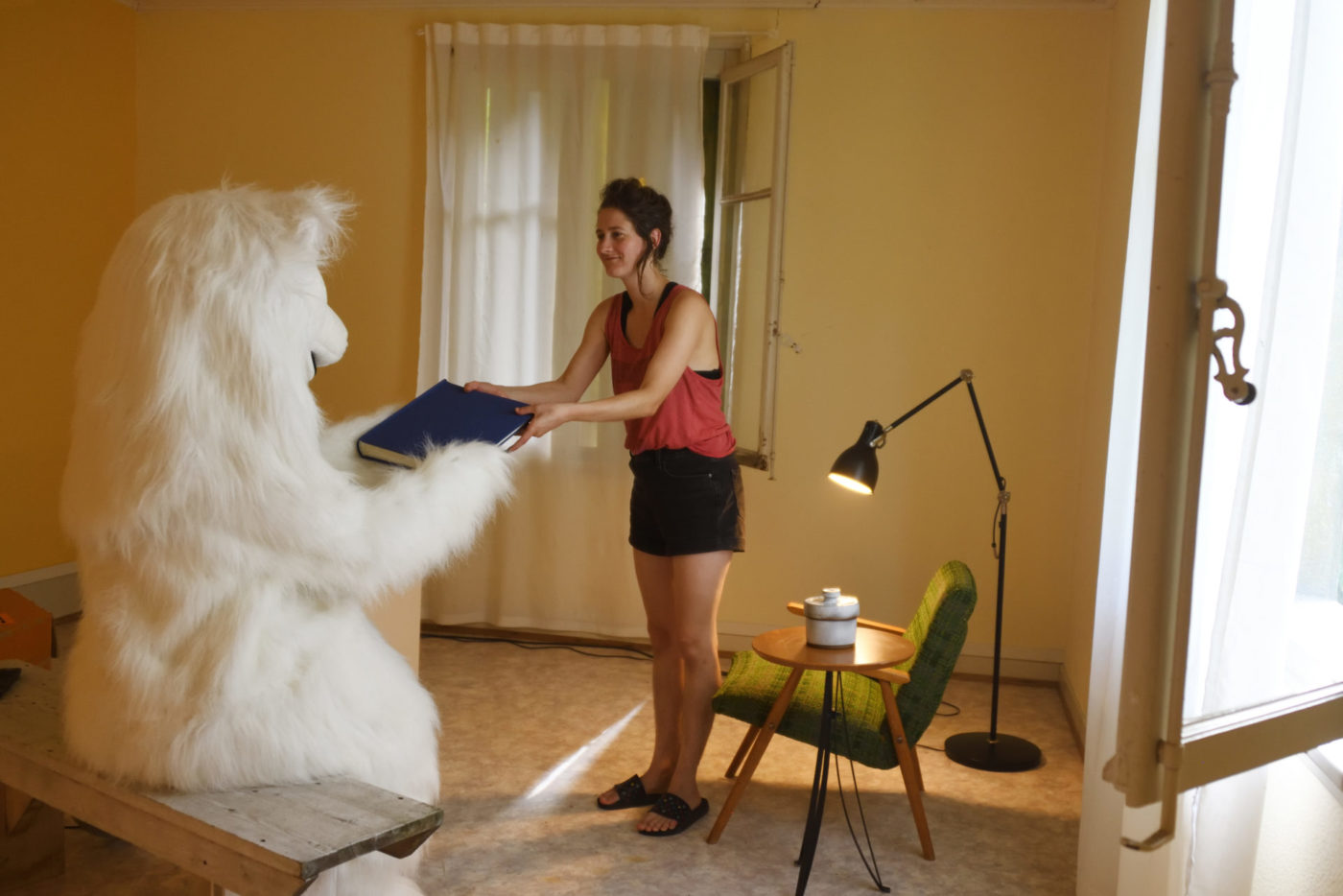 The artist Camilla Parini stands in a little room. The room is lighted and cosy, with a table, an armchair and a living room lamp. She hands a big black book to a person disguised as a polar bear.