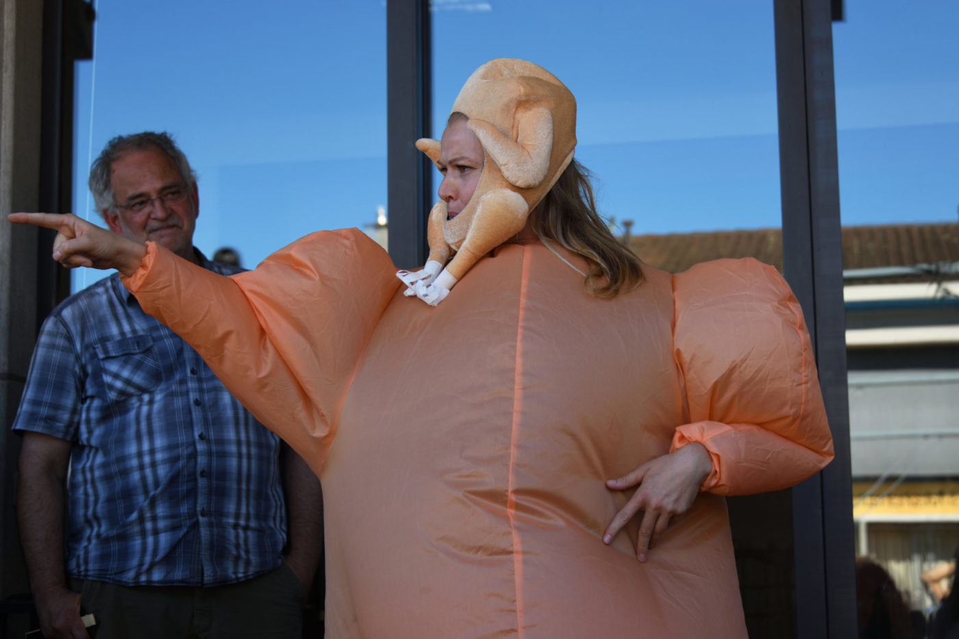 The artist Danae Dario performs outside before a window. She is disguised in an orange inflated fabric. Around her face is fixed an artificial cooked chicken.