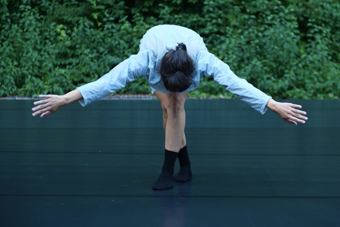 The dancer Marion Baeriswyl stands on a scene in the forest. She takes a bow. Her whole upperbody is directed toward the floor, arms opened.