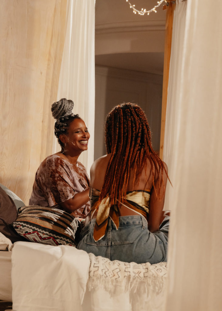 The artist Lucile Saada Choquet sits on a bed and looks on her right at a person that came to participate to her performance, to talk about her experience as a racialised person.