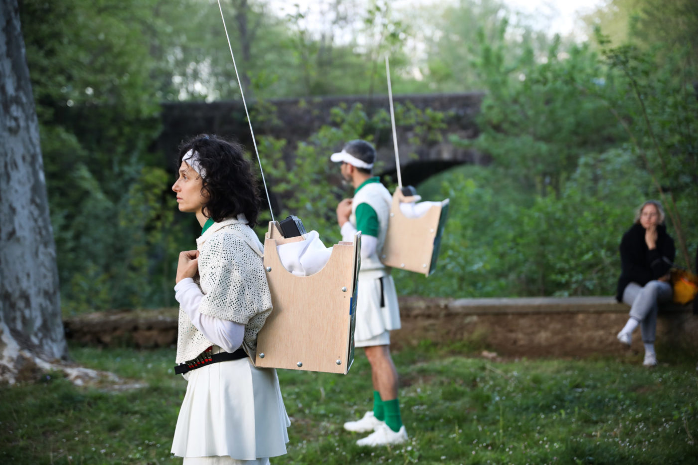 The scene takes place outdoors, in a grassy field. Two people are holding bags made of wood with radio antennas sticking out.