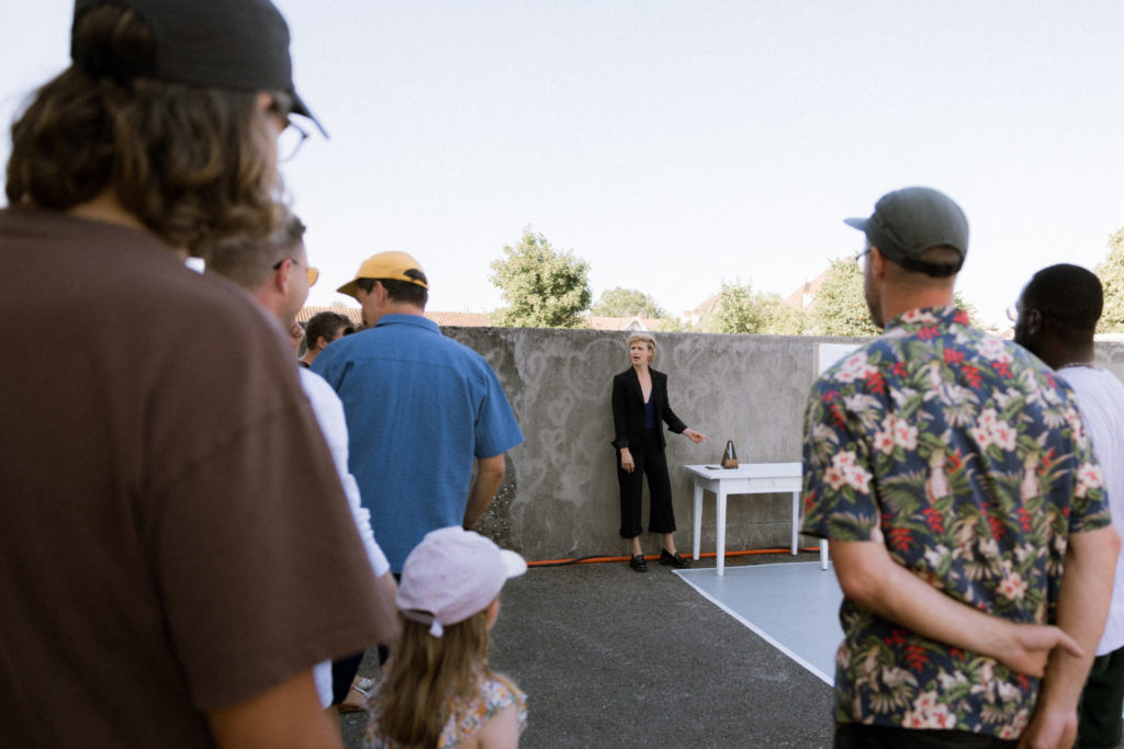The public is seen from behind in la cour des marchandises. The public looks in the direction of the artist Salomé Mooij, who stands in the center at the back, mimicking a movement of stumble.