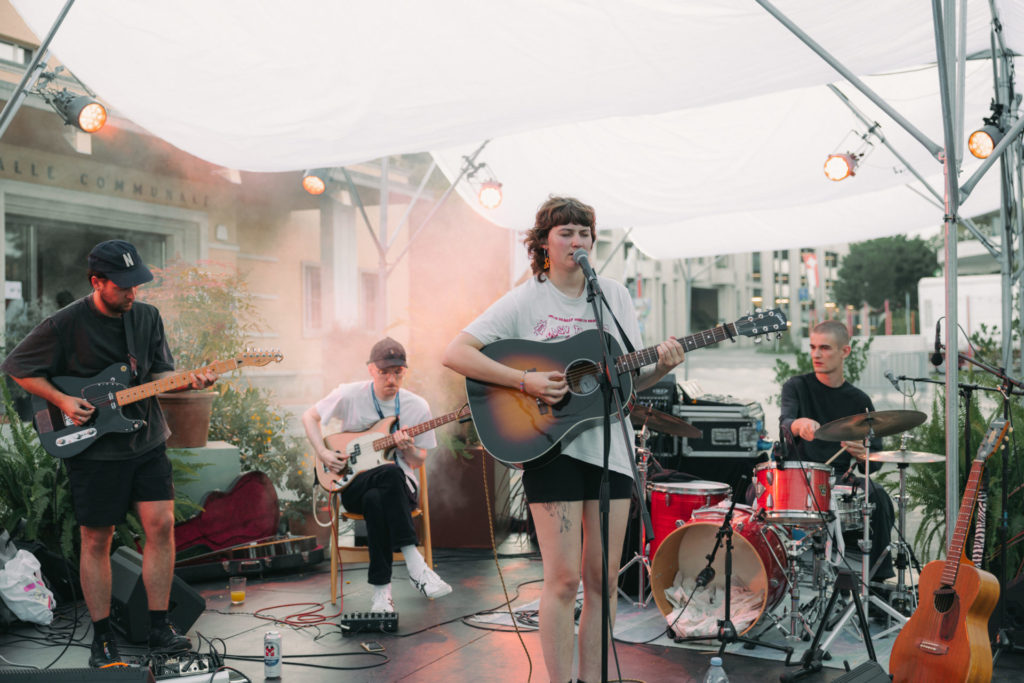 We can see a music band on a scene. The singer clair my flair stands in the front and sings. Behind her, we can discover three musicians surrounding her.