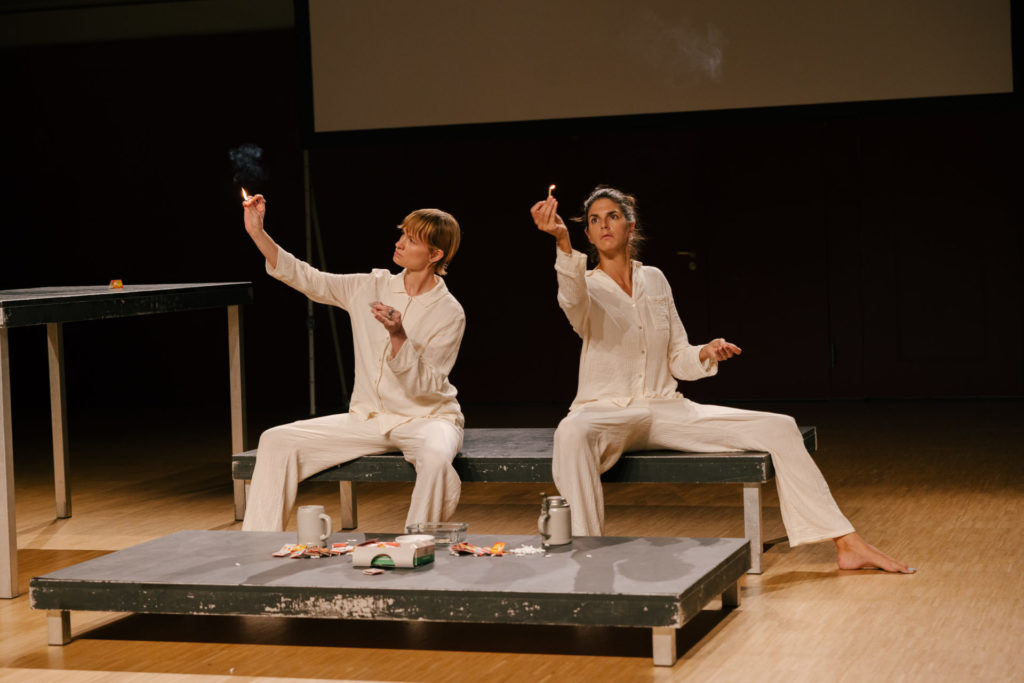 The two comedians sit on a bank. They were pyjamas and have in one hand a lighted match, that they are looking at.