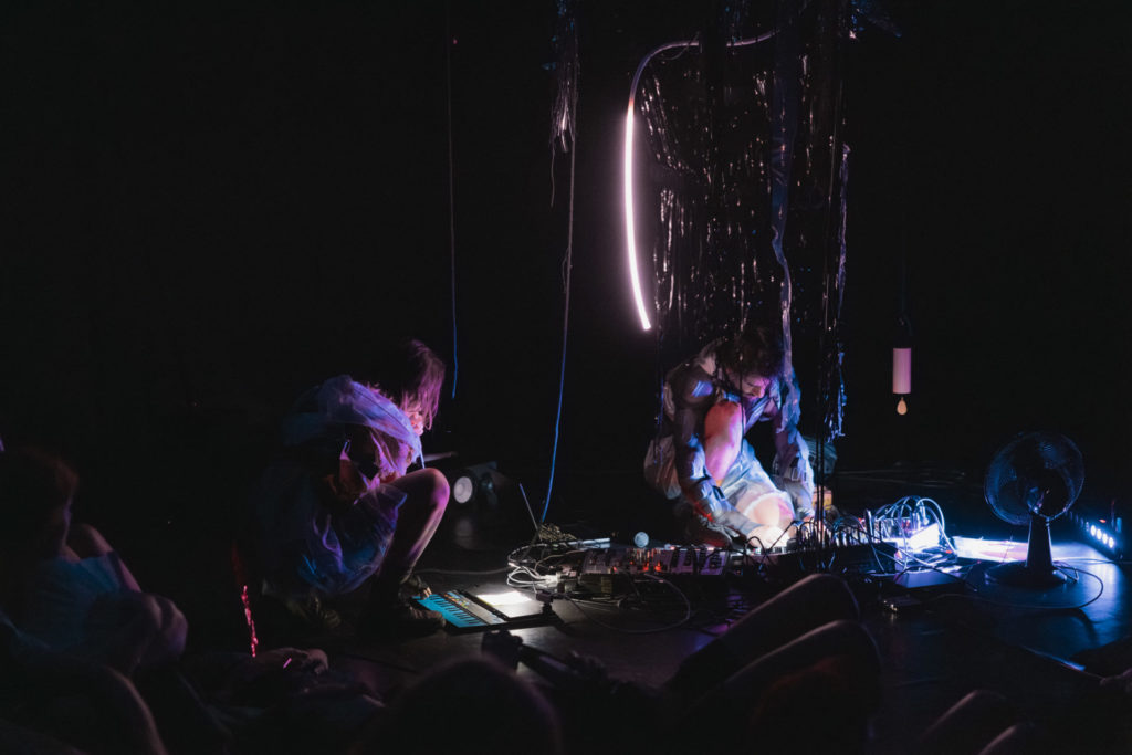 We can see the artist Francesca Sproccati on the left on the floor, creating sounds on a big synthesizer. On her left, a musician does the same. It is obscure in the room, only purple neons lights illuminate the room.