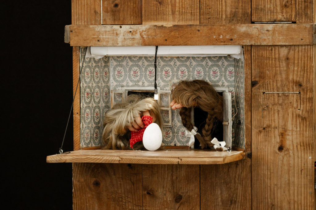 There is an opening in a big wooden box. From this opening we can see two hands wearing wigs and facing each other. They look at an egg in the center and get out of two little doors.