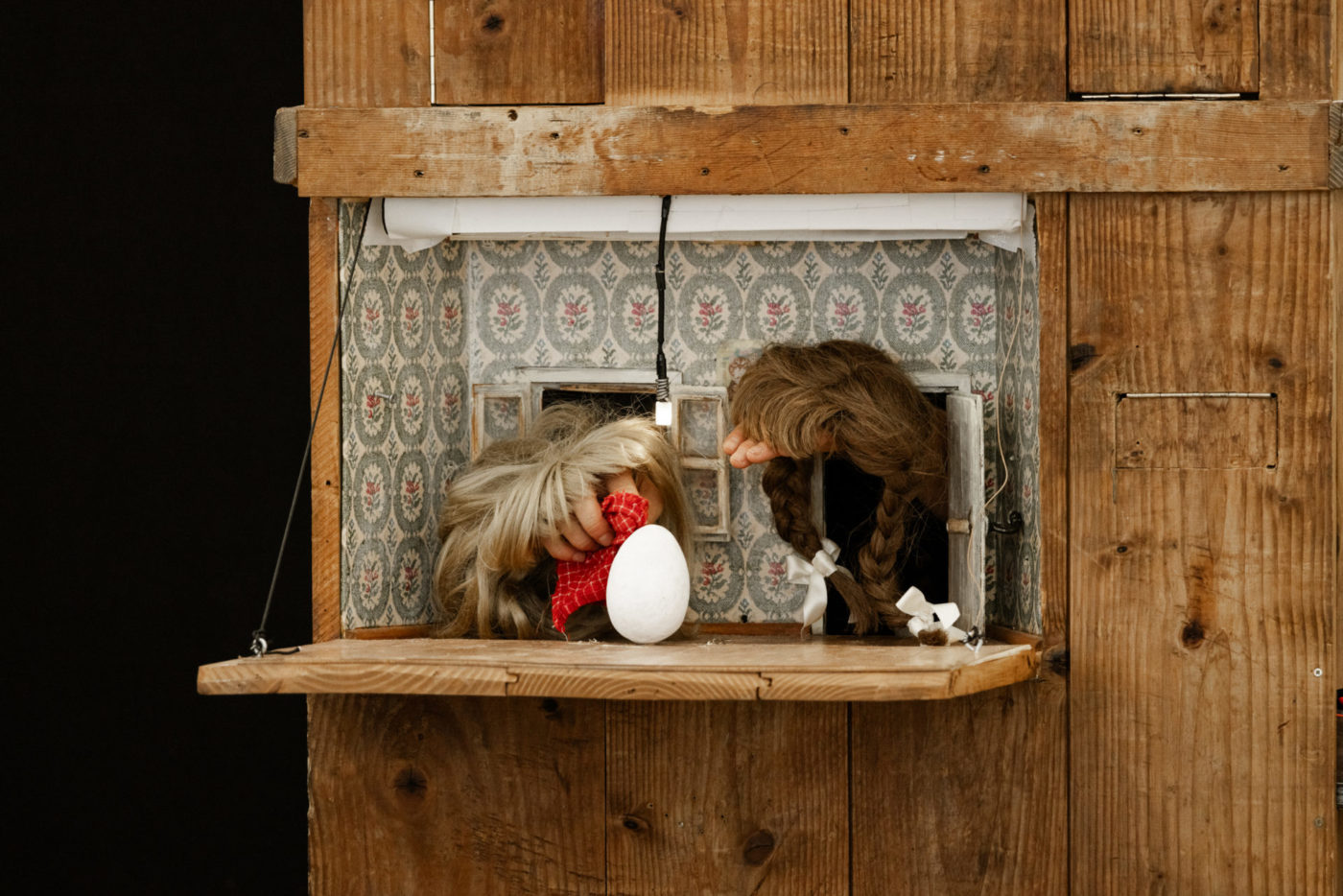There is an opening in a big wooden box. From this opening we can see two hands wearing wigs and facing each other. They look at an egg in the center and get out of two little doors.