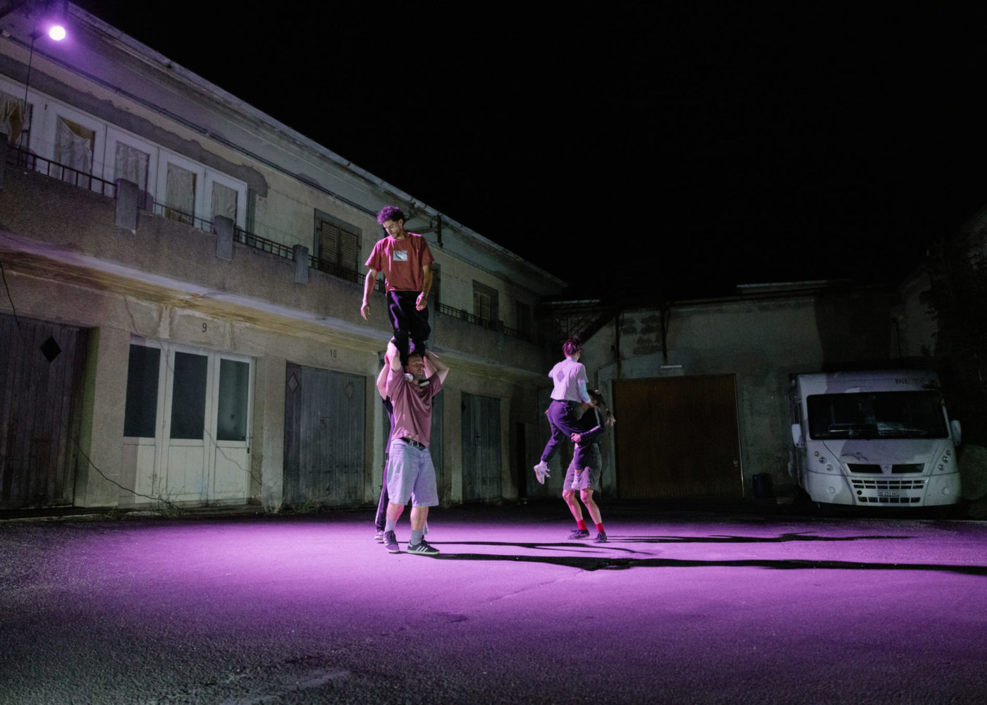 Five circus artists are evolving in a courtyard. A first group of two support one circus artist who stands upright on their hands and shoulders. In the second group one circus artist is raised by another to prepare for a new figure.