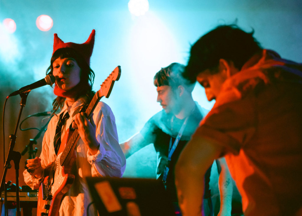 On the right we see the singer of the band 24 KIARA. She wears a red crochet hat with two extremities, resembling a cat. Behind her we see two other musicians, one playing the keys and the other the guitar.
