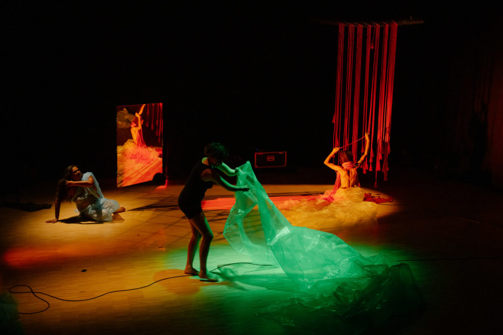 The three artists occupy different areas of the scene. On the left on the floor, Living Smile Vidya sits in her blue plastic costume. At the center, Meret Landolt grasps a wide green plastic surface with her short arms . On the right, Nina Langensand is naked, seen from behind, and rolls up around her neck red ropes that cover her progressively.