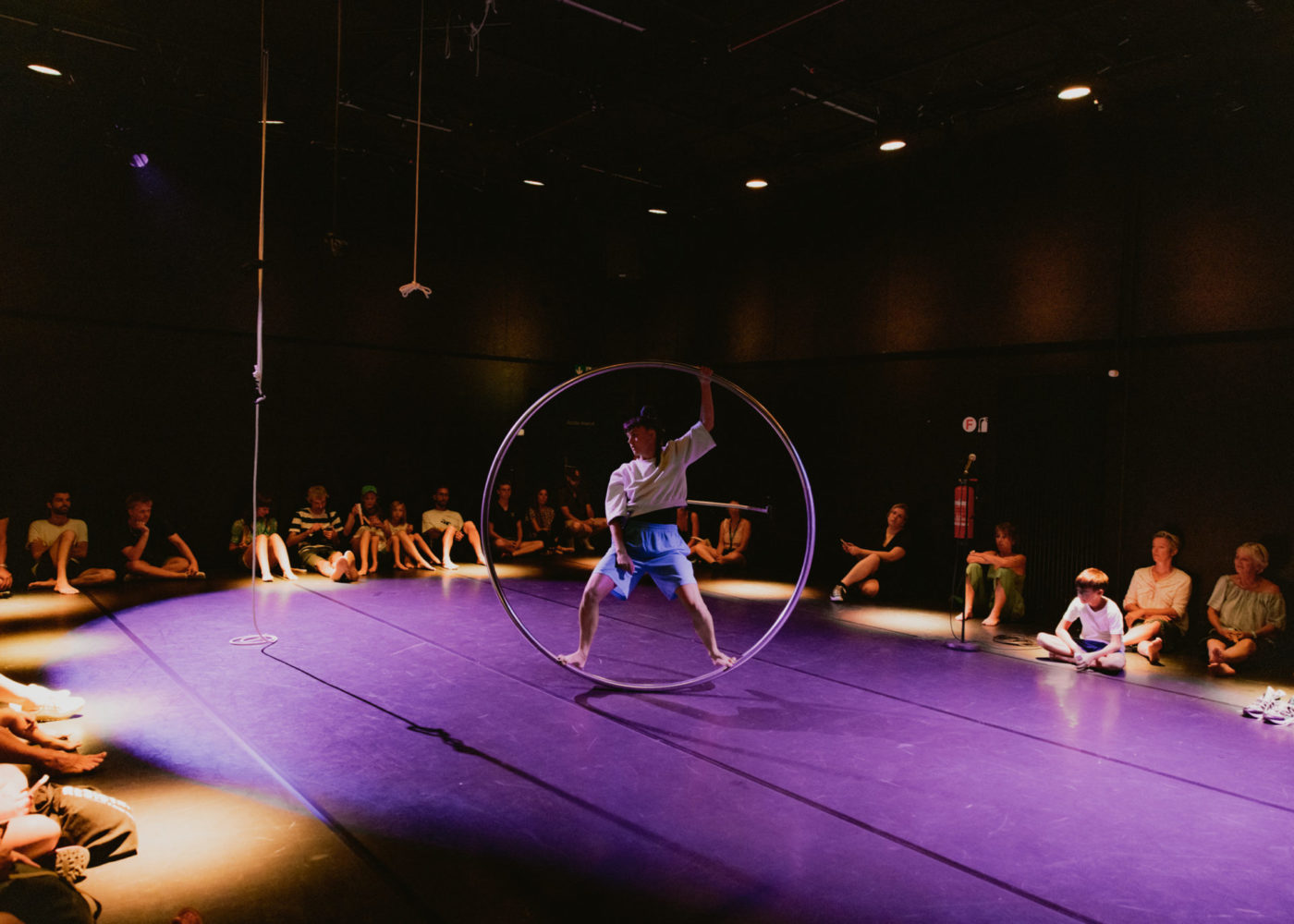 The circus artist lili parson piguet stands on a wheel. She supports herself with one hand on the wheel and her two feet on the wheel. The public watches and surrounds her.