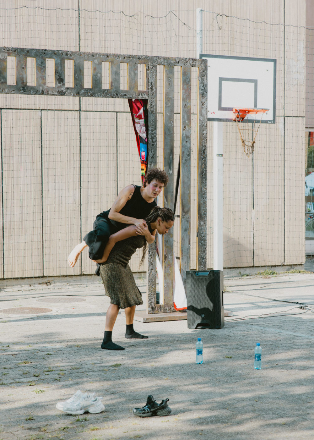 One of the two dancers supports the other on her back. They have removed their shoes. The performance occurs next to a basketball court et we notice a speaker in the back.