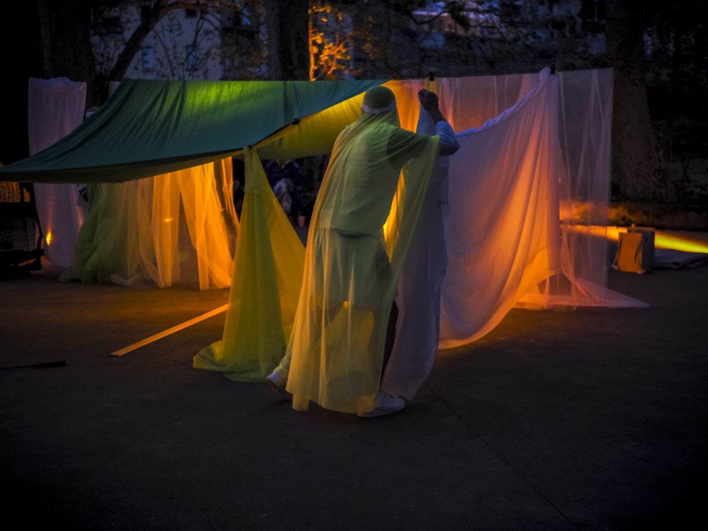 Large fabrics are spread out outdoors. An actor is handling these large fabrics illuminated with different colors.