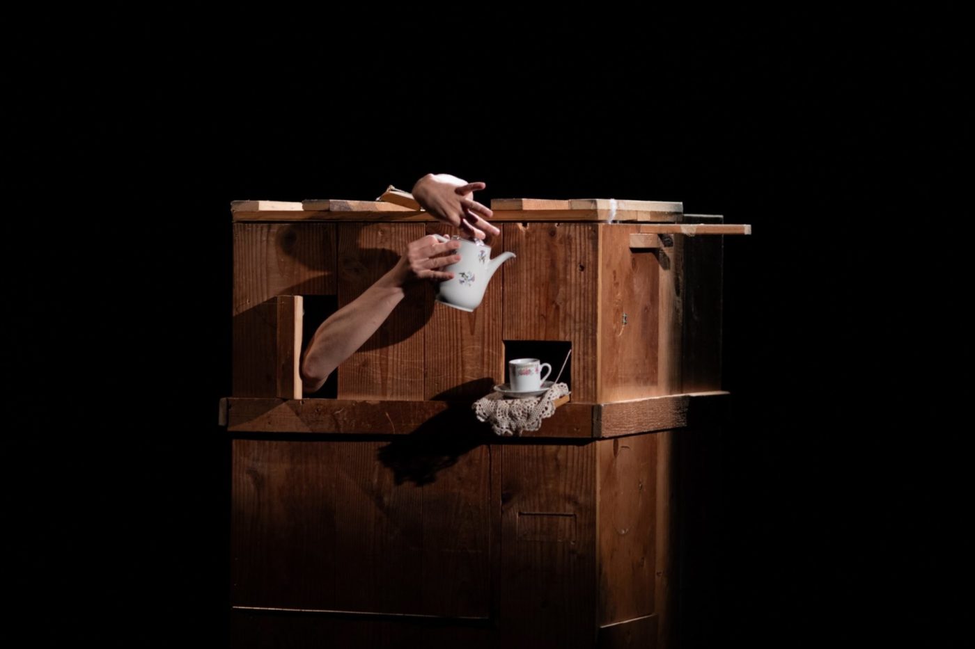 From a wooden box, two hands emerge holding a teapot to pour a cup of tea.