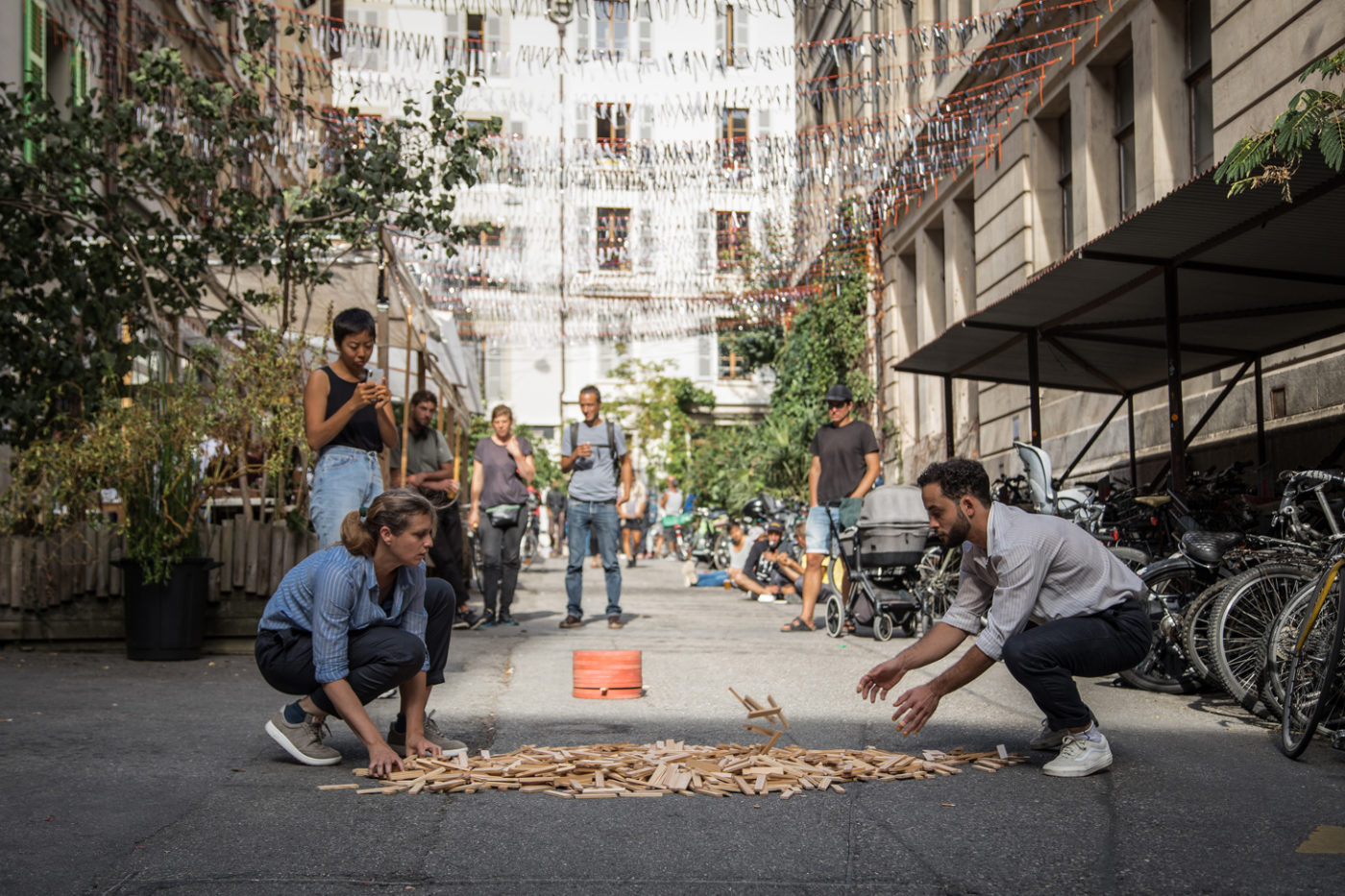Two people are playing with KAPLA® blocks spread out on the ground, in a public space. People are watching them.