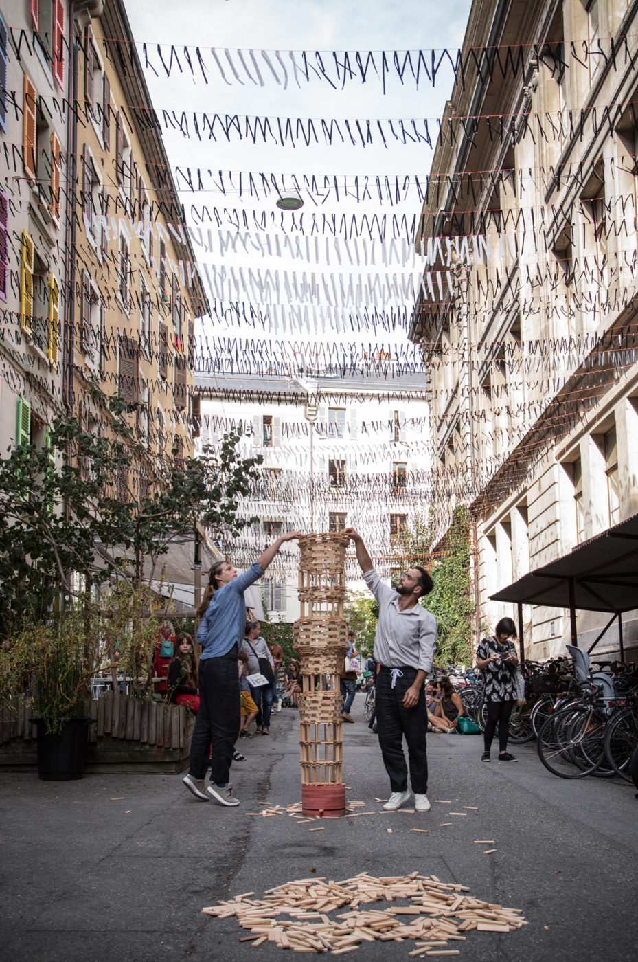 Two people are building a tower in a public space. The tower is made of KAPLA® blocks.