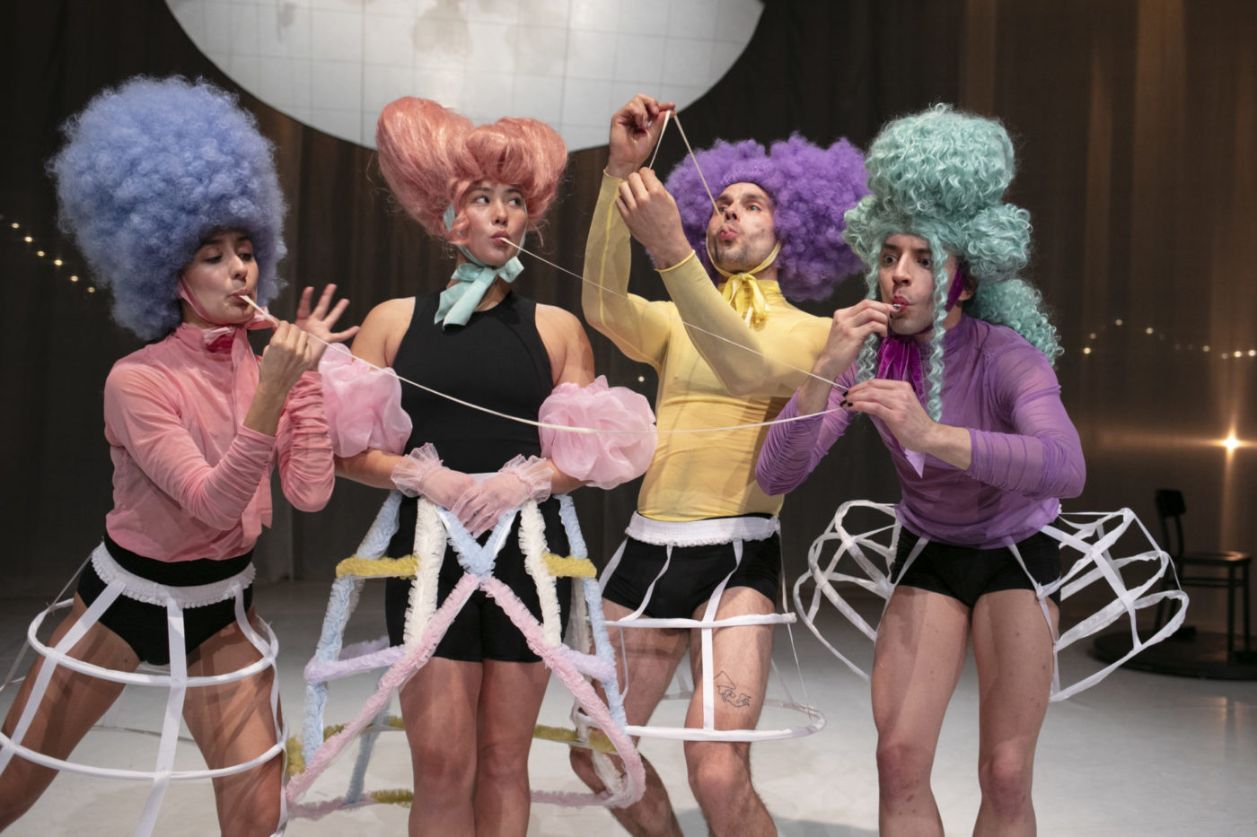 Four dancers are on stage. They are wearing wigs in different colors: blue, orange, purple, and green. Their colorful tops match their wigs. They are also wearing long checkered skirts, reminiscent of what women wore under their dresses in the 18th century.