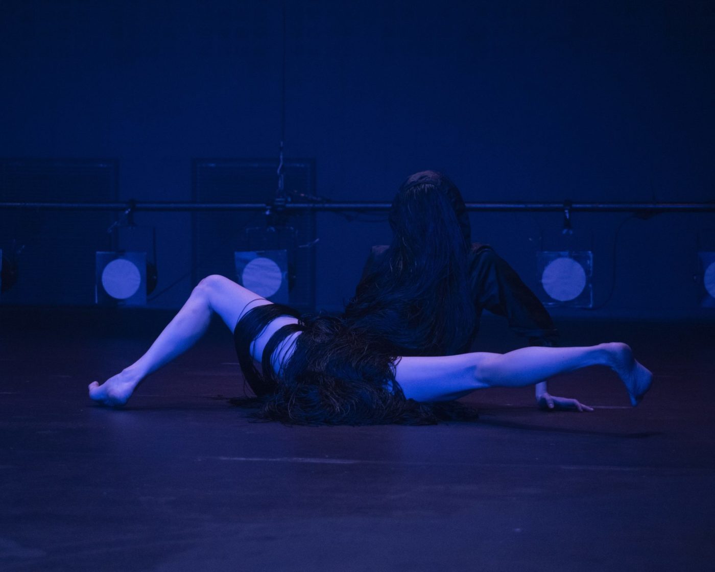 Marion Zurbach is dressed in black, in a dark room. Her legs are bare. She is doing the splits. She is wearing a black wig. It gives the impression of a strange animal shape.