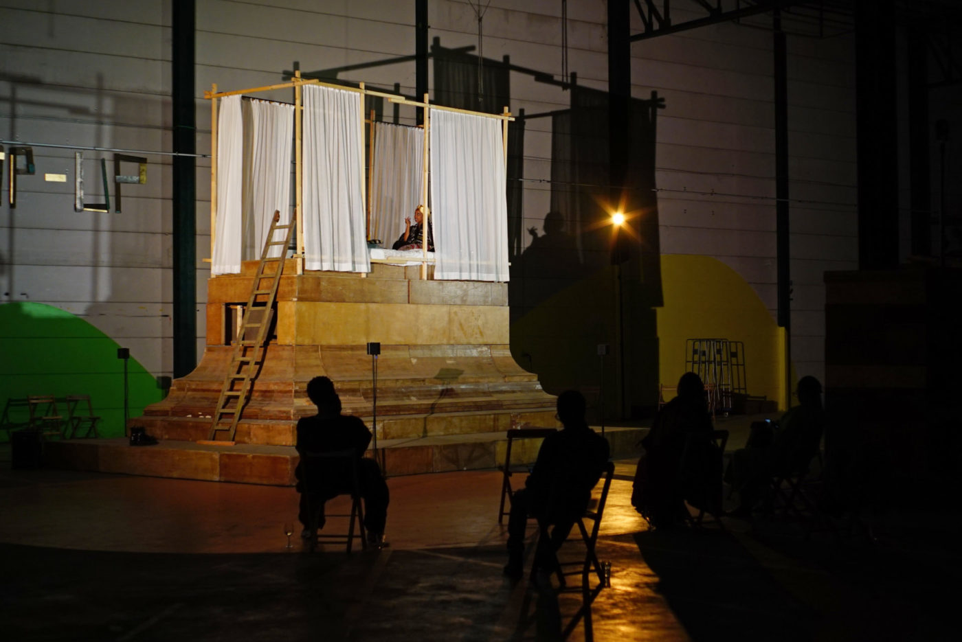 Dans une salle de théâtre, il y a un lit surélevé qui baigne dans la lumière. Le reste de la pièce est dans l'obscurité. Le lit est entouré de rideaux. Lucile Saada Choquet se trouve dans le lit.