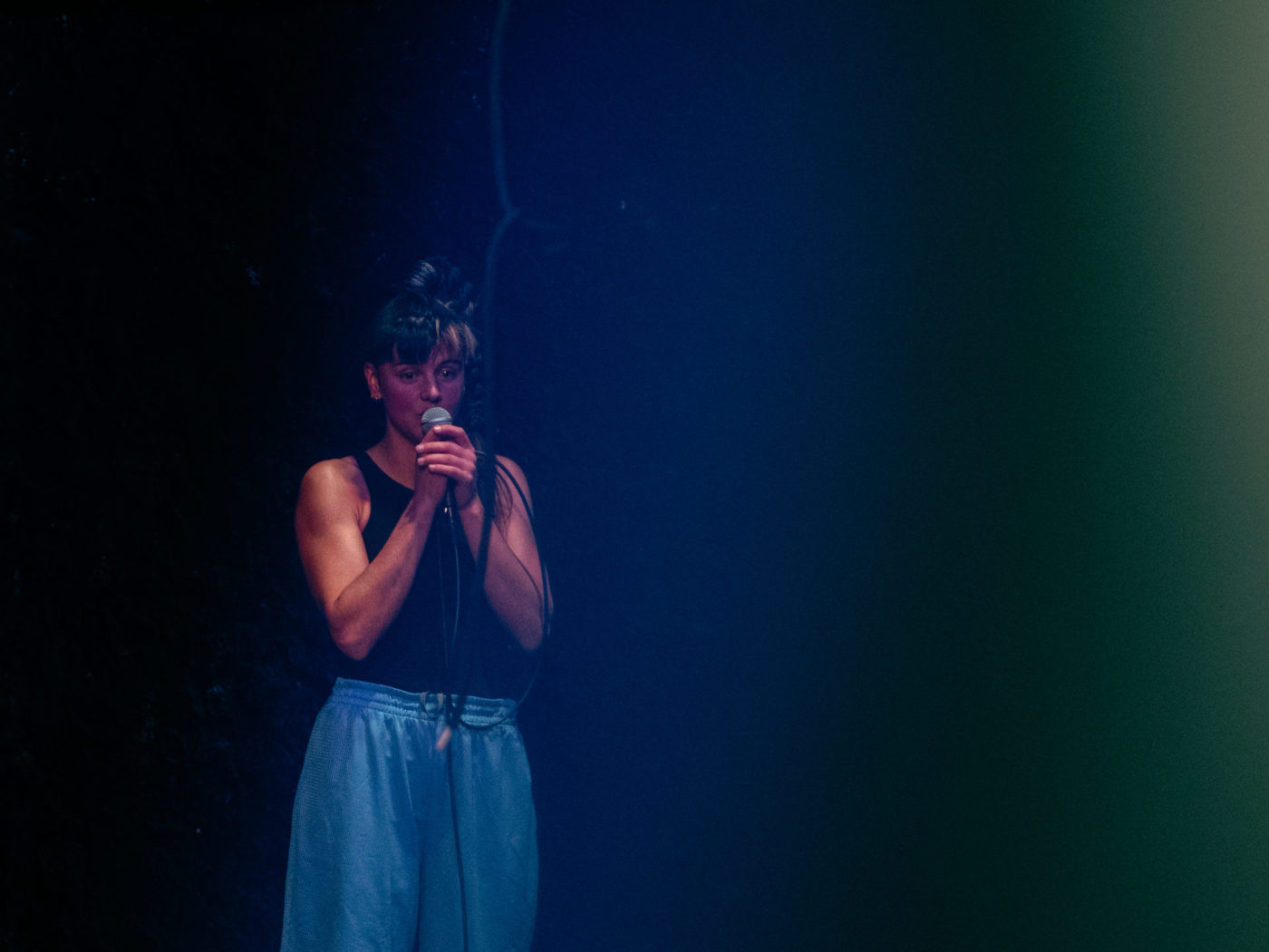 lili parson piguet is standing, wearing blue shorts. She is holding a microphone in her hands. She looks like she is telling a story.