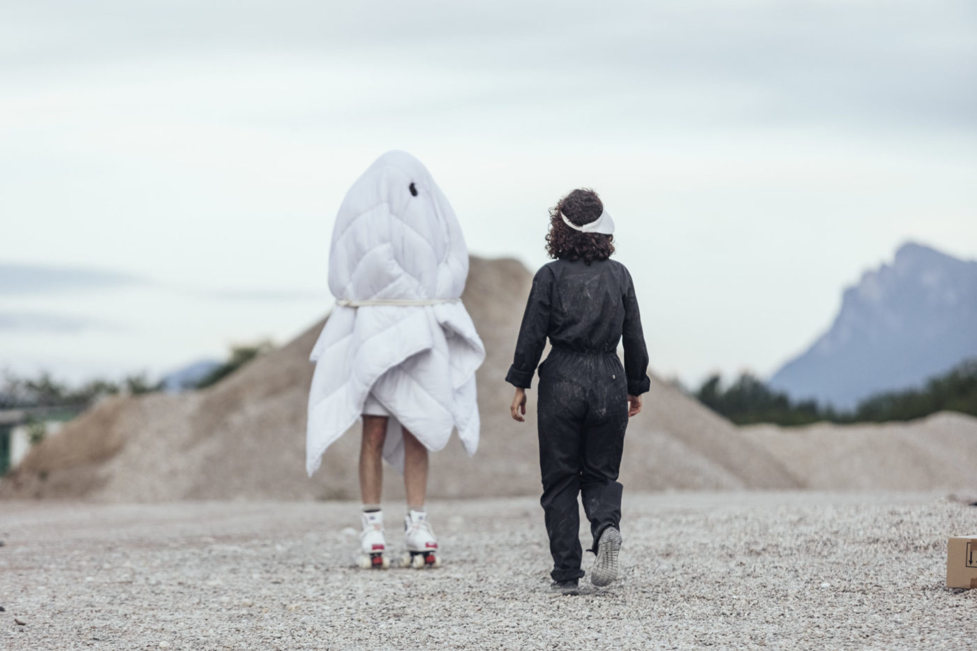 Two people are outdoors. The landscape is a gravel pit. The people are walking. On the right, the person is wearing a black fabric jumpsuit. On the left, the person is wrapped in a large costume made of white sheets.
