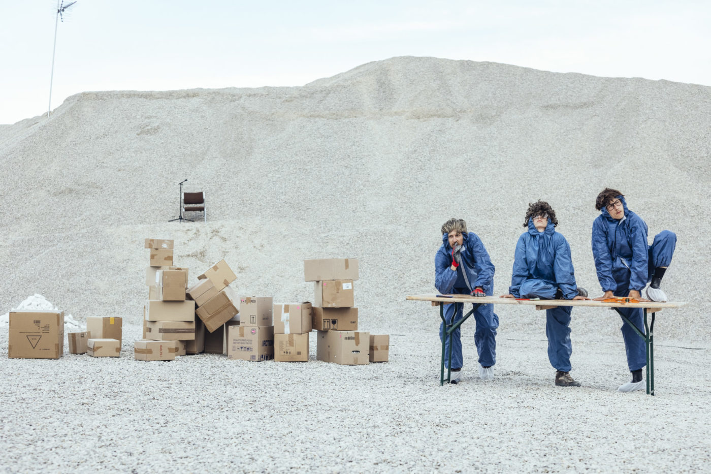Three actors are wearing blue fabric jumpsuits. They have brown wigs and fabric shoe covers. Their outfits look a bit like hospital uniforms. On the left, there are stacked cardboard boxes, and behind them, a huge pile of gravel.