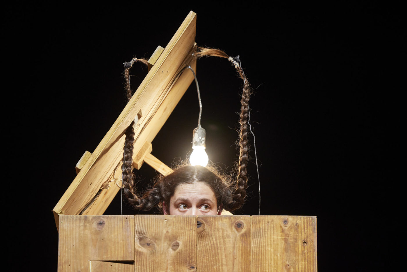 Annina Mosimann is inside a wooden box. She is sticking her head out of the box, up to the middle of her face. Her hair, braided into two plaits, stands upright above her head. A large lightbulb hangs from the lid of the box, just above her head.
