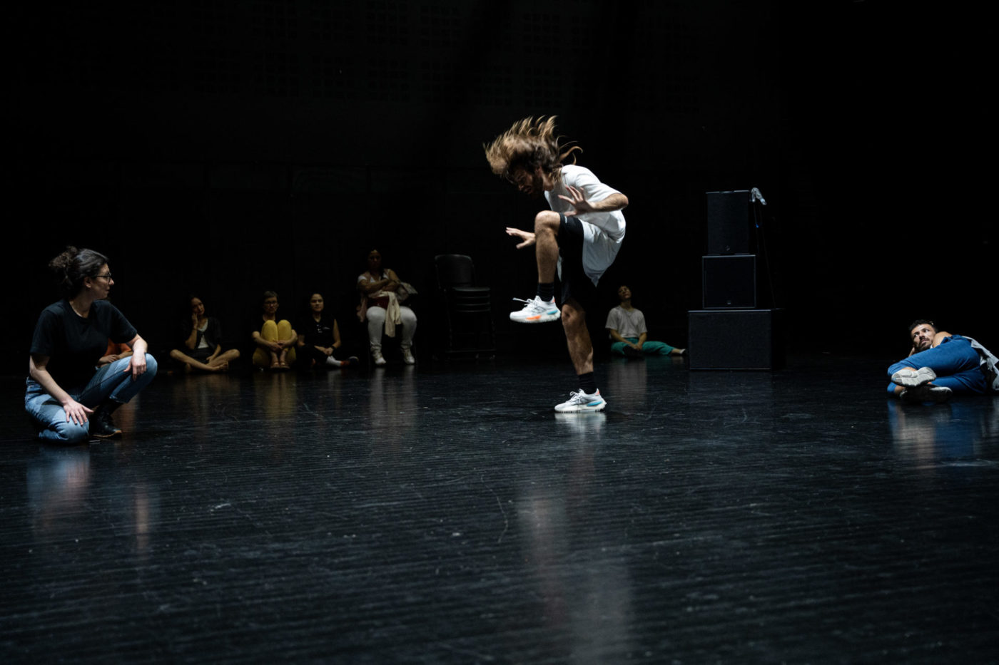 Ametonyo Silva danse dans une salle noire, il a l'air de rebondir. Ses cheveux sont ébouriffés et il a un genou levé. Du public est autour de lui dans la salle.