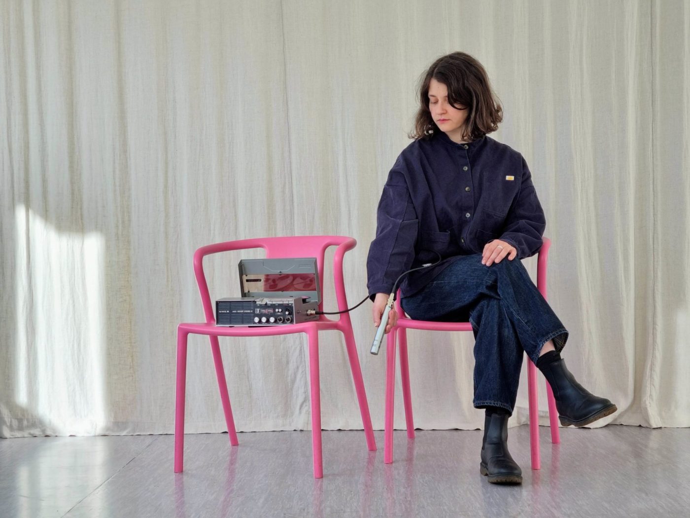 Annina Polivka is sitting on a red chair. She is holding a microphone in her hand. The microphone is connected to a machine that produces sound. The machine is also placed on a pink chair next to the artist.